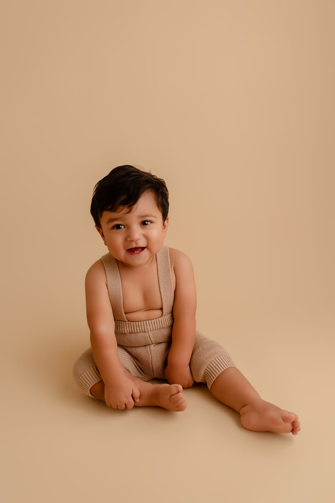 Baby in cream overalls sitting against a cream backdrop